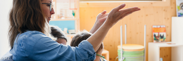 Eine erwachsene Person sitzt mit zwei Kindern in einem hellen Raum auf einem Sofa. Die Person trägt eine blaue Bluse, spricht oder erklärt etwas und macht dabei eine Gebärde beziehungsweise eine deutende Handbewegung. Eines der Kinder sitzt direkt neben ihr und blickt in dieselbe Richtung. Im Hintergrund sind Spielmaterialien, Bücher und helle Holzwände zu sehen.