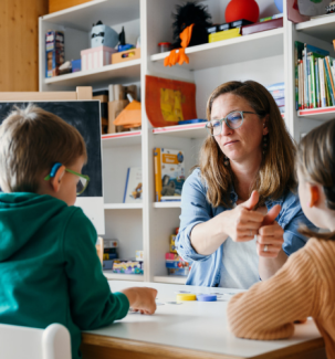 Eine erwachsene Person sitzt mit zwei Kindern an einem Tisch und spielt ein Spiel. Dazu wird eine Gebärde zeigt.