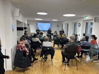 Blick in einen hellen Veranstaltungsraum mit zahlreichen sitzenden Teilnehmenden. Vorne halten zwei Frauen einen Vortrag zum Thema psychische Erkrankungen. Eine Gebärdensprachdolmetscherin übersetzt den Vortrag in Gebärdensprache. Im Hintergrund ist eine Präsentation auf einem Bildschirm zu sehen.